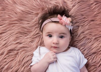 Baby girl wearing pink headband lying on pink blanket looking at the camera for her newborn baby photoshoot in Northampton