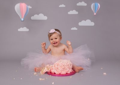 One year old baby girl sitting in front of pink cake on grey background for baby cake smash photoshoot