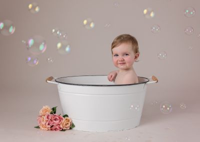 Baby girl in tub with bubbles and flowers on the floor for first birthday cake smash photography session