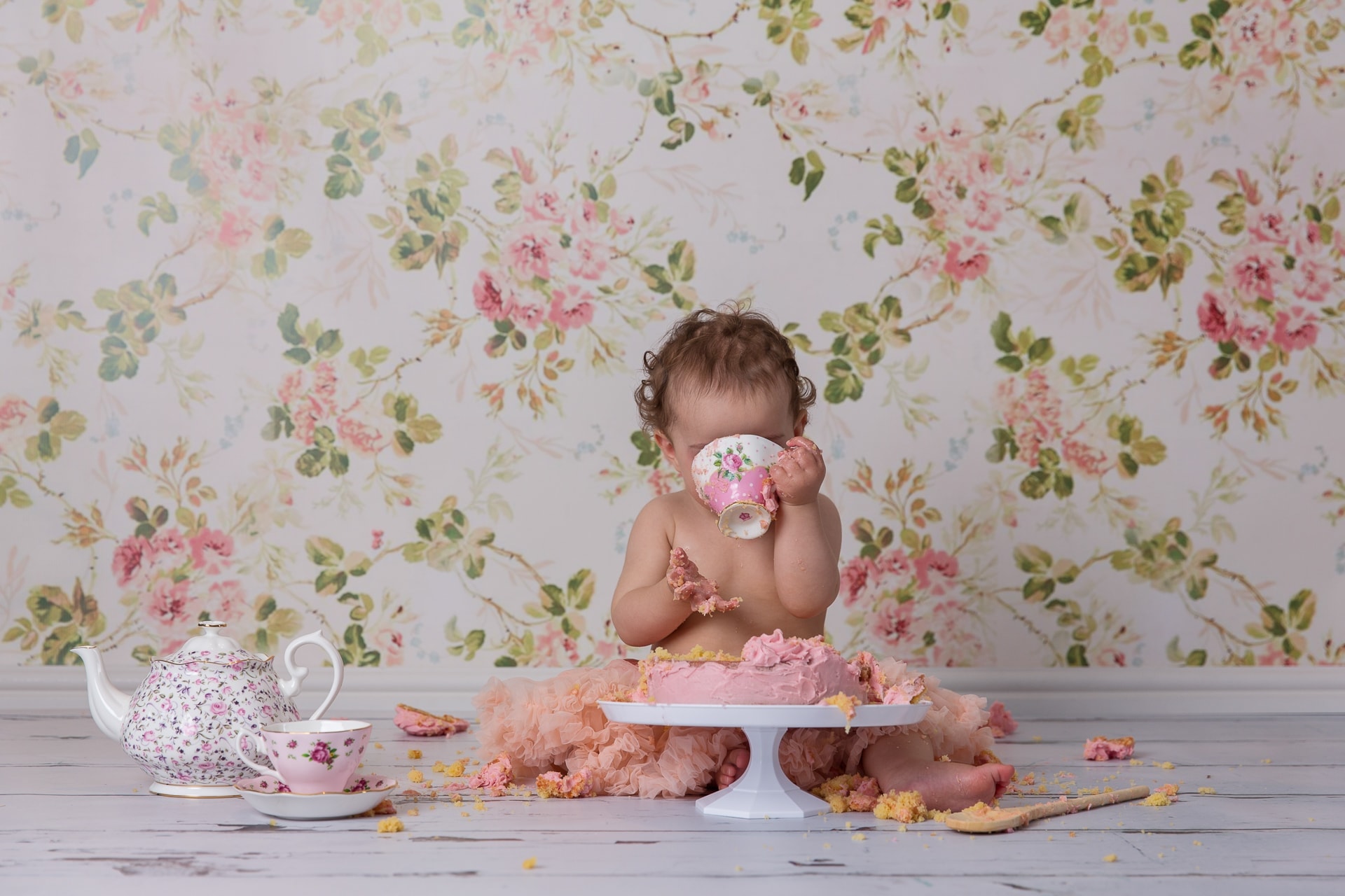 Baby cake smash photography showing one year old girl sipping from tea cup in front of smashed cake