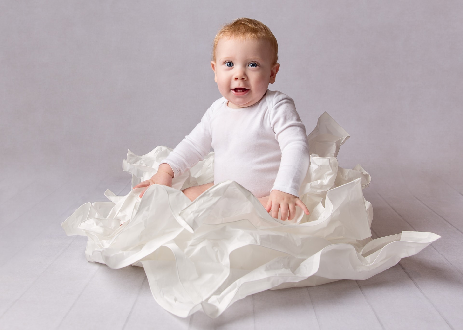 Six month old baby boy wearing white vest sitting in white paper on baby photoshoot