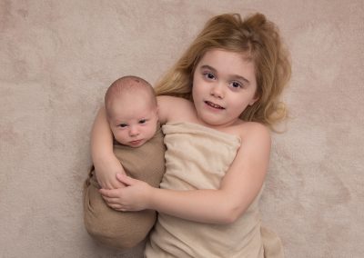 Baby boy and big sister looking at the camera on beige background for newborn photoshoot in Northampton